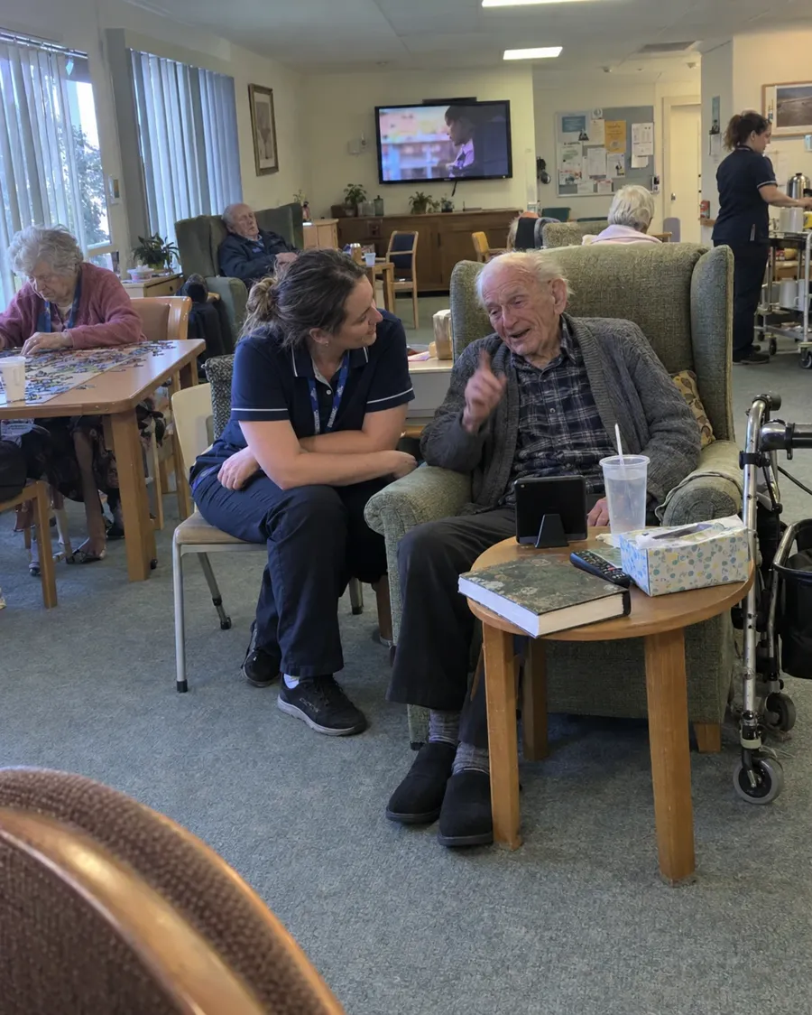 An older man in an armchair telling a story to a phone on the table beside him, while a carer listens